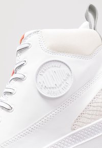 Gros plan d'une basket montante blanche avec un patch rond du logo de la marque Palladium et des lacets blancs avec des coutures noires.