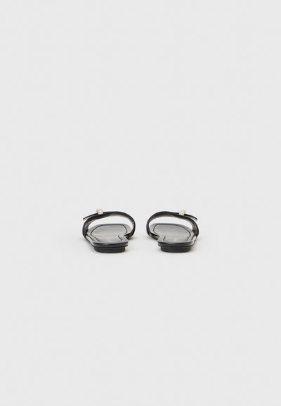 Paire de sandales plates noires avec boucle pour l'orteil et design minimaliste, vues de face sur un fond gris clair.