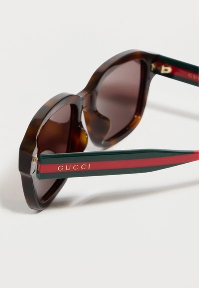 Lunettes de soleil rondes en écaille de tortue avec verres marron, dotées de branches vertes et rouges et d'un logo Gucci doré sur le côté.