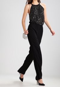 Combinaison noire avec un haut à col halter en sequins, design à jambes larges, et un sac à main argenté. Mannequin portant des talons hauts noirs.