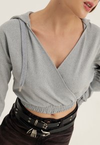 Cropped hoodie grigio realizzato in morbido tessuto, con scollo a V, cappuccio con lacci e vita elastica. Abbinato a una cintura nera.