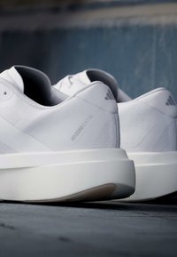 Ζευγάρι λευκά παπούτσια για τρέξιμο Adidas Adizero Evo SL με παχιά σόλα πάνω σε σκοτεινή επιφάνεια και θολό φόντο.