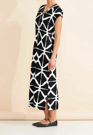 Robe sans manches noir et blanc avec un décolleté en V, présentant un motif géométrique et des fentes sur les côtés. Associée à des chaussures plates noires.