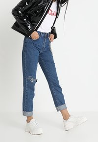 Czarna błyszcząca kurtka puchowa, biała koszulka z czerwonym napisem, niebieskie jeansy o średnim stanie z postarzanymi detalami oraz białe masywne sneakersy.