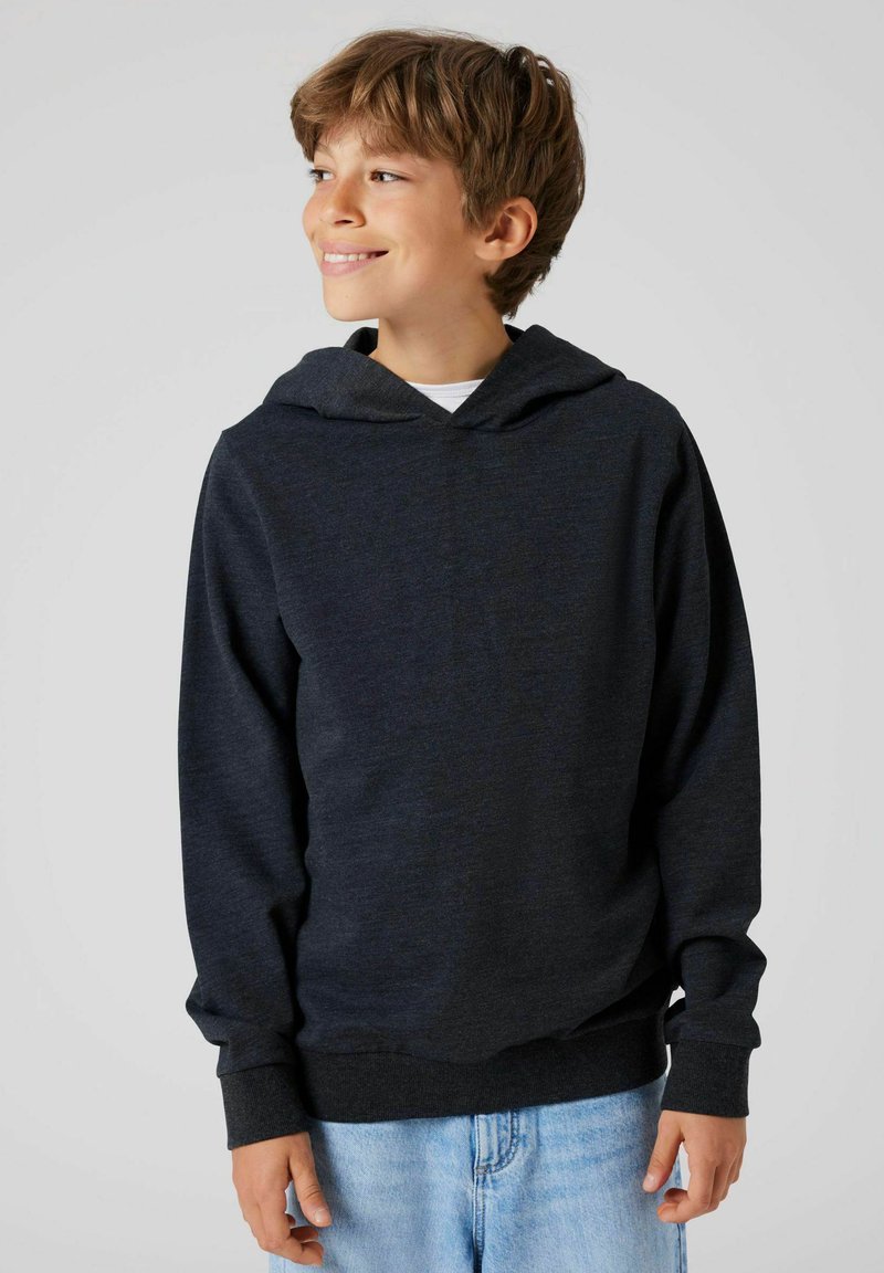 Lächelnder Junge mit kurzem, hellbraunem Haar trägt einen dunkelgrauen Kapuzenpullover und hellblaue Jeans. Er schaut zur Seite vor einem schlichten grauen Hintergrund.