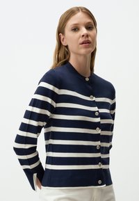Cardigan a righe navy e bianche con maniche lunghe, scollatura rotonda e bottoni argentati sul davanti. Tessuto morbido, design aderente.