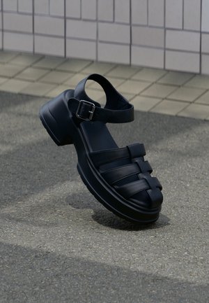 Schwarze Leder-Sandale mit geschlossenem Zeh, Knöchelriemen, Metallschnalle, dicker Sohle, liegend auf grauem Pflaster nahe einer gefliesten Wand.