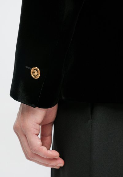 Μαύρο βελούδινο σακάκι με χρυσό διακοσμητικό κουμπί που φέρει σχέδιο λιονταριού στη μανσέτα. Ομαλή υφή και διακριτή ραμμένη σιλουέτα.