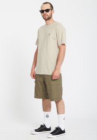 Beżowa koszulka bawełniana z małym logo, połączona z oliwkowymi szortami cargo. Dodatki to czarne okulary przeciwsłoneczne i czarne sneakersy z białymi skarpetami.