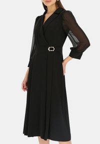 Czarna sukienka kopertowa z przewiewnymi, półprzezroczystymi rękawami, dopasowaną talią podkreśloną ozdobną klamrą i plisowaną spódnicą.