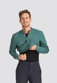 Green, long-sleeve button-up shirt with a mandarin collar, featuring a textured fabric and a relaxed fit, worn over a black top.