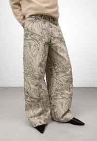 Osoba w szerokich, beżowych spodniach w zielony marmurkowy wzór, w beżowym, puszystym swetrze i czarnych, spiczastych butach, stojąca na szarym podłodze.