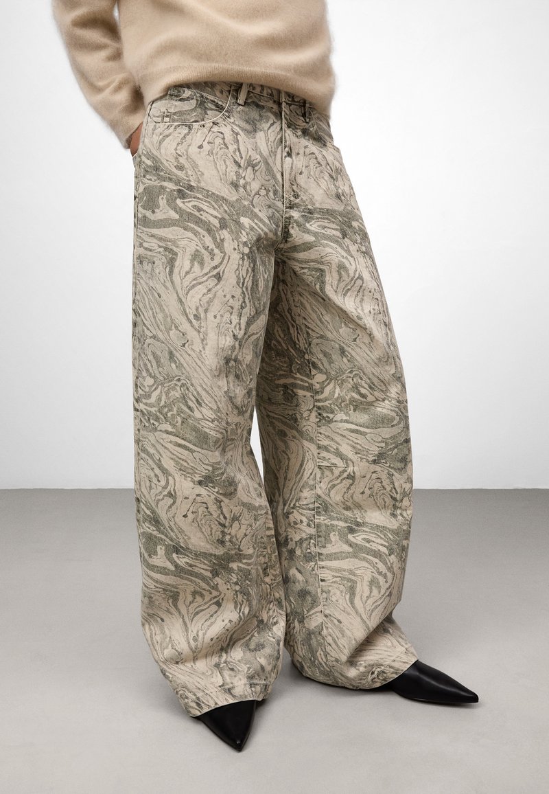 Osoba w szerokich, beżowych spodniach w zielony marmurkowy wzór, w beżowym, puszystym swetrze i czarnych, spiczastych butach, stojąca na szarym podłodze.