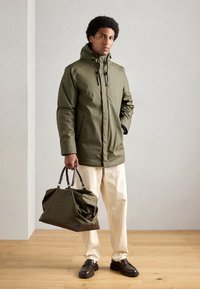 Grøn vandtæt parka med hætte og snoretræk, parret med cremefarvede bukser og mørke loafers, som holder en stor olivengrøn taske med brune detaljer.