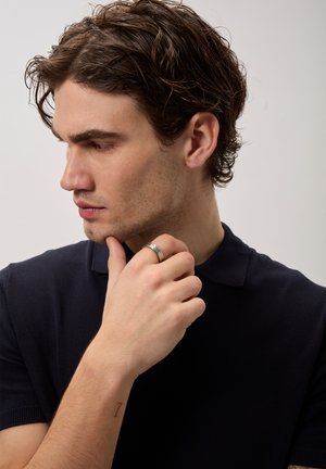 Młody mężczyzna z ciemnymi falowanymi włosami, ubrany w czarną koszulę, dotykający lewą ręką podbródka i noszący srebrny pierścionek na palcu.