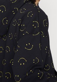 Black hoodie made of cotton blend, featuring a repeating pattern of yellow smiley faces. Hooded design with a soft texture.