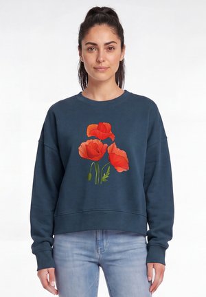 Femme aux cheveux foncés en queue de cheval, portant un sweat-shirt bleu foncé avec des coquelicots rouges imprimés sur le devant et un jean bleu clair.