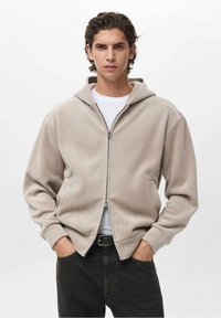 Felpa con zip beige realizzata in tessuto testurizzato, dotata di cappuccio, due tasche laterali e polsini a costine. Indossata sopra una maglietta bianca e jeans scuri.