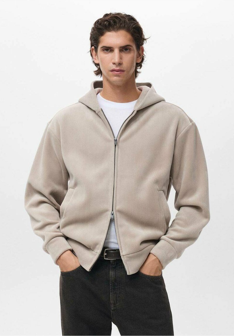 Felpa con zip beige realizzata in tessuto testurizzato, dotata di cappuccio, due tasche laterali e polsini a costine. Indossata sopra una maglietta bianca e jeans scuri.