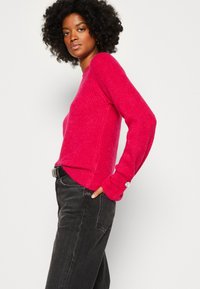 Fuchsia gebreide trui met een gestructureerde oppervlakte, lange mouwen en een subtiele knoopdetail aan de manchetten, gecombineerd met donkere high-waisted spijkerbroeken.