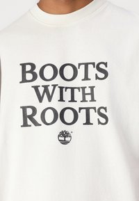 Krém színű pamut pulóver, amelyen vastag fekete betűkkel szerepel a "BOOTS WITH ROOTS" felirat, valamint egy kis logó található, amelyen egy körben egy fa látható.