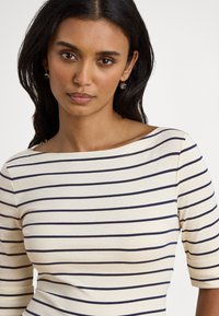 Γυναίκα με σκούρα μαλλιά που φοράει κρεμ πουκάμισο με ναυτικό οριζόντιο ριγέ και μικρά κρεμαστά σκουλαρίκια, κοιτάζοντας μπροστά, με απλό φόντο.