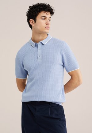 Młody mężczyzna z ciemnymi kręconymi włosami, ubrany w jasnoniebieską teksturowaną koszulkę polo z krótkim rękawem i ciemne spodnie, patrzący na bok.