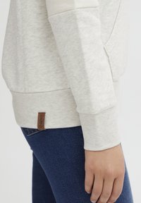 Felpa beige chiaro con tessuto testurizzato, polsini a coste e un'etichetta laterale con il logo "oxmo", abbinata a jeans blu scuro.