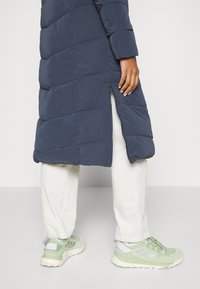 Marineblå pufferjakke med quiltet mønster, sidelukning, parret med lyse brede bukser og grønne sneakers med detaljer og snørebånd.