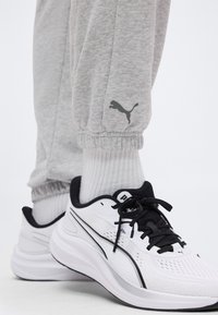 Λευκά και μαύρα αθλητικά παπούτσια Puma, φορεμένα με λευκές ριμπ κάλτσες και γκρι φόρμες με λογότυπο Puma στο τελείωμα.