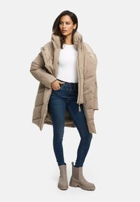 Manteau long beige rembourré avec col haut, fermeture éclair et poches latérales. Porté sur une chemise blanche et un jean bleu. Bottines beiges.