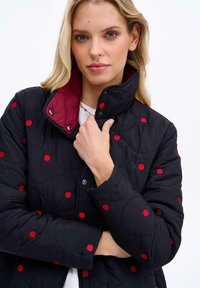 Prešívaná čierna bunda s výšivkou červenej jablka, marhuľovým golierom, cvokovými uzávermi a jemnou textúrou. Má priliehavý strih a jemný lesk.