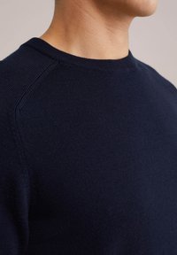 Temno modri pleteni pulover s krožno izrezom, gladko teksturo in raglan rokavi. Lahkoten material, zasnovan za udobje in prileganje.