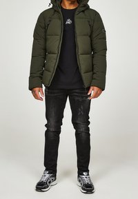 Mand iført en olivengrøn polstret jakke, sort grafisk t-shirt, sorte slidte jeans og sort-hvide sneakers på en ensfarvet baggrund.
