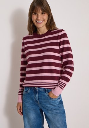 Femme souriante portant un pull rayé bordeaux et rose avec un jean bleu, debout avec une main dans la poche sur un fond uni.