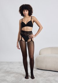 Conjunto de lingerie preto com um bralette e cuecas de cintura alta. Meias de rede pretas transparentes completam o visual. Textura suave e design ajustado.