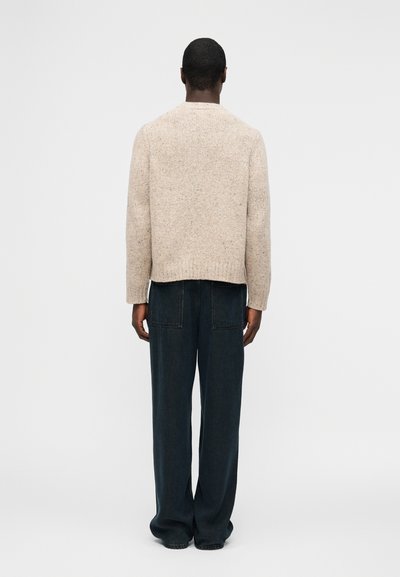 Άνδρας που στέκεται πλάτη προς το μέρος μας, φοράει μπεζ πλεκτό πουλόβερ και φαρδιά σκουρό μπλε παντελόνια με μεγάλες πίσω τσέπες, σε απλό φόντο.