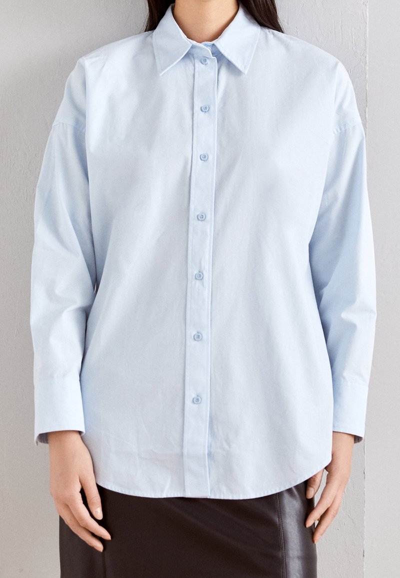 Helles blaues Hemd mit Kragen, langen Ärmeln und abgerundetem Saum. Aus einem glatten Stoff mit einem schlichten Design gefertigt.