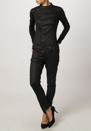 Femme portant une veste noire cintrée avec une fermeture éclair asymétrique, un pantalon noir slim et des talons aiguilles noirs, debout, jambes croisées.