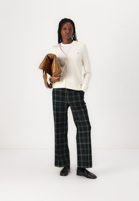 Look stratificato con un cardigan crema, camicia bianca, pantaloni a quadri scuri con accenti verdi e una borsa in pelle verniciata marrone.