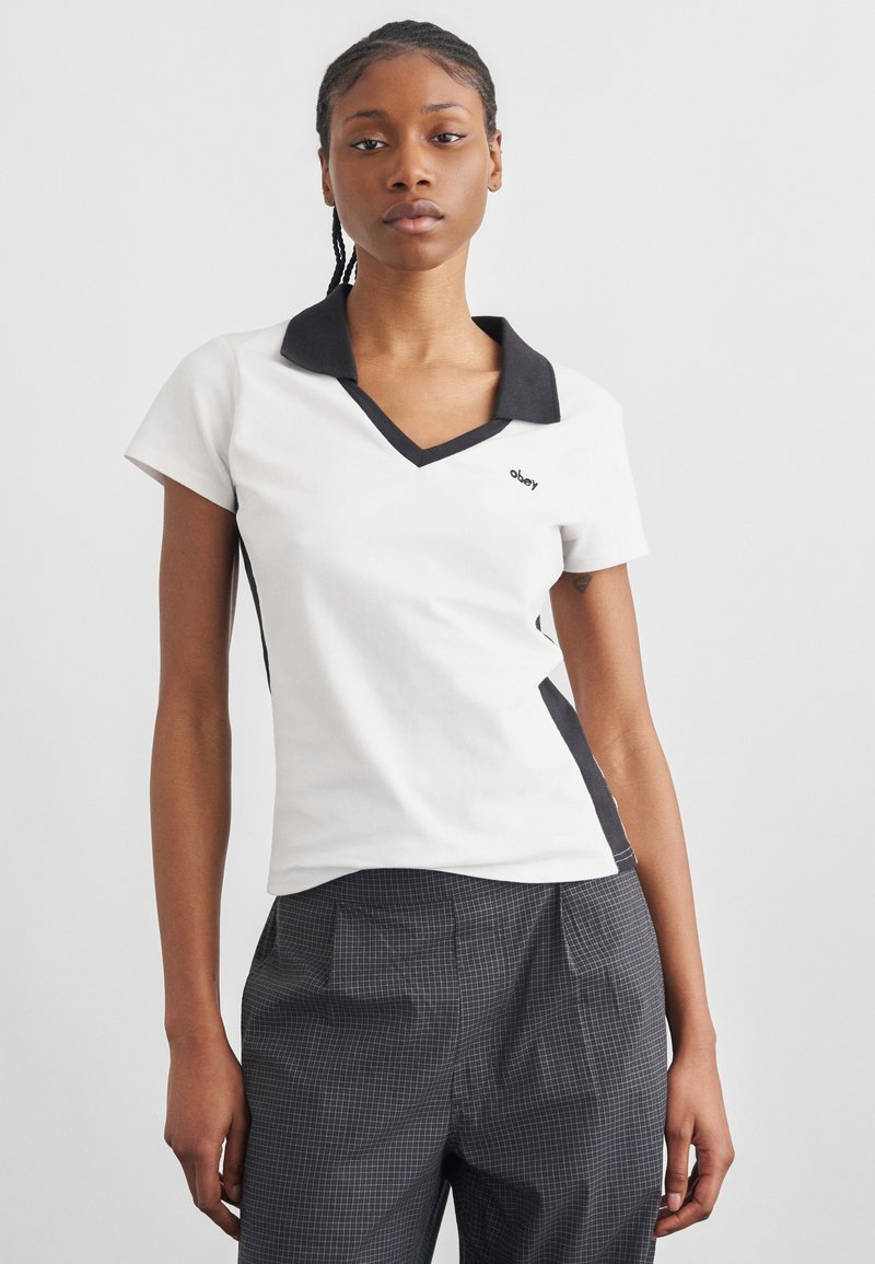 Νεαρή γυναίκα που φοράει λευκό κοντομάνικο τοπ με μαύρο γιακά και πλευρικές πινελιές, και ψηλόμεσο καρό παντελόνι, στέκεται μπροστά σε απλό φόντο.