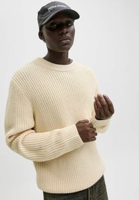 Pull beige en tricot avec un motif texturé et larges poignets côtelés, associé à une casquette noire avec un logo blanc.