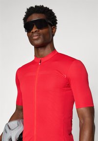 Maillot de cyclisme rouge ajusté à manches courtes, texture lisse, avec une fermeture éclair frontale ; porté avec un casque de vélo gris et des lunettes de soleil sombres.