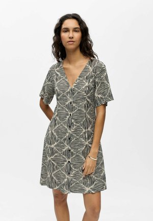 Mladá žena v krátkych rukávoch, šaty s výstrihom do V a čierno-béžovým abstraktným geometrickým vzorom, stojaca na jednoliatom bielom pozadí.