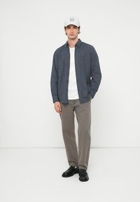 Tengerészkék kockás gombos ing, fehér póló, szürke nadrág és fekete cipő. A modell fehér logós sapkát visel. Egyszínű háttér.