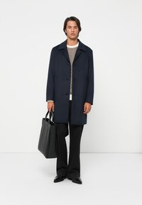 Cappotto navy con colletto classico, cinque bottoni neri, indossato sopra un maglione a fantasia. Il modello tiene una borsa a tracolla nera, abbinata a pantaloni neri.