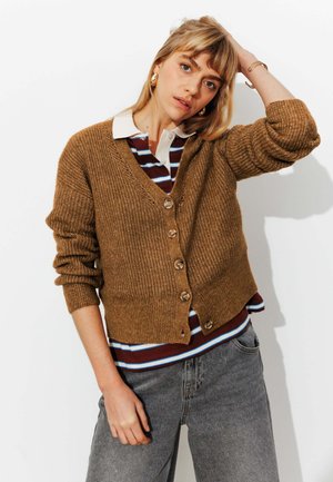 Jeune femme aux cheveux blonds porte un cardigan marron boutonné sur un polo rayé et un jean gris, posant avec un bras levé.