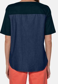 Blusa a maniche corte con parte superiore verde scuro e parte inferiore in denim blu scuro, caratterizzata da un orlo arrotondato e dettagli di cucitura visibili.