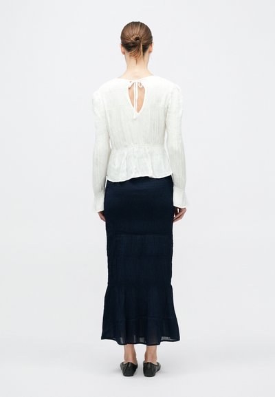 Nő kontyba fogott hajjal, fehér, hátul megkötős részletekkel ellátott hosszú ujjú blúzban és sötétkék, boka alá érő szoknyában, fekete balerinacipőben.