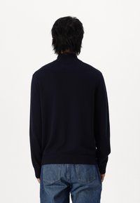 Maglione blu navy con collo alto, maniche lunghe e una texture liscia. Orlo e polsini a coste, mostrato da dietro. Abbinato a jeans azzurri chiari.